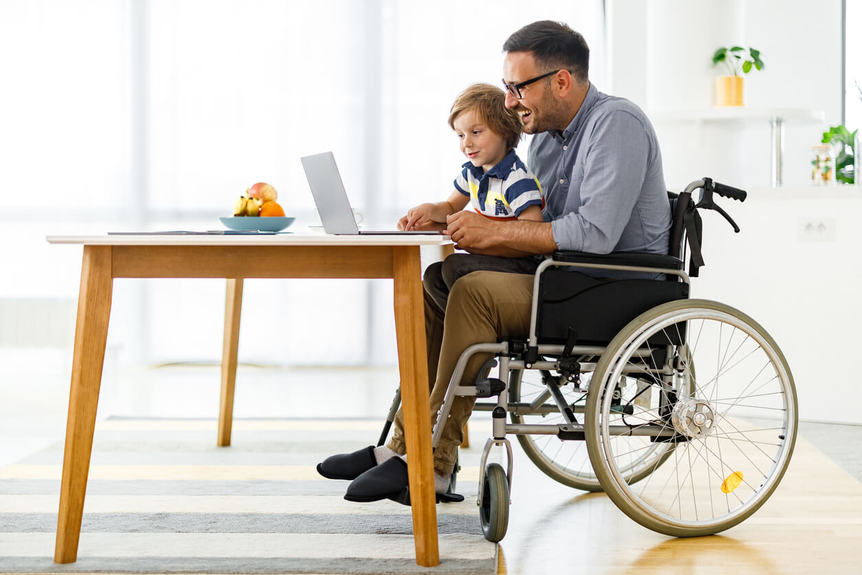man in wheel chair at a desk with child on his lap in front of laptop